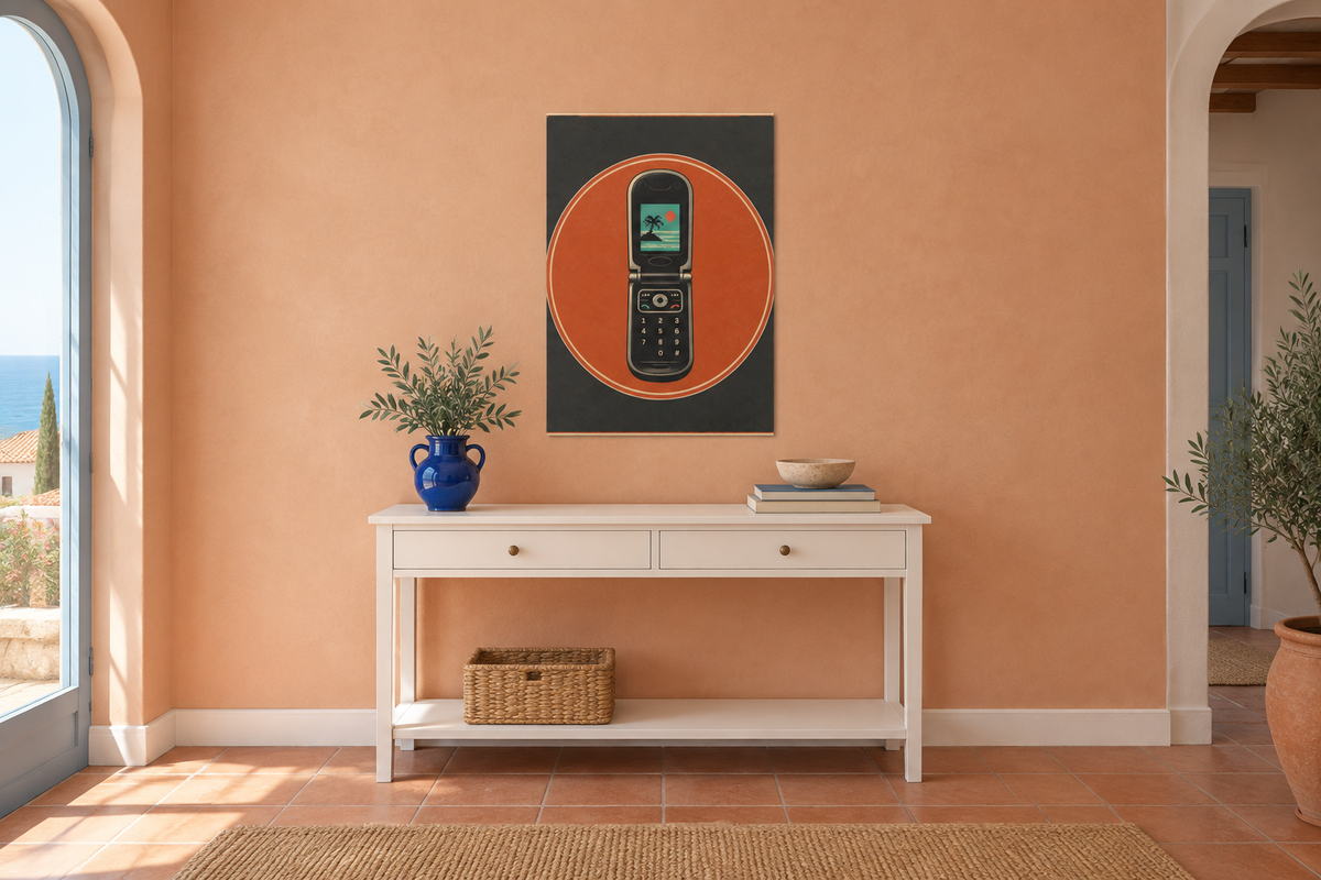 A framed wall art print featuring a retro flip phone inside an orange oval on a dark background, displayed above a white console table in a warm peach-colored room.
